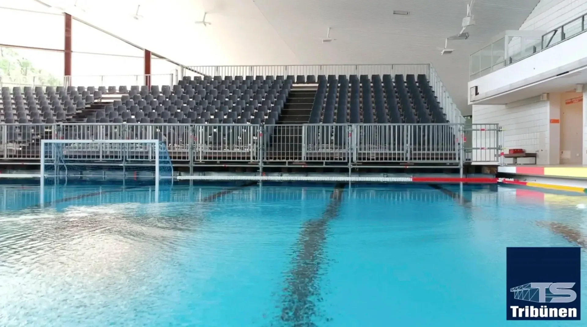 Hallenbad mit leeren Tribünen. Blaues Wasser, weiße Geländer, rote und gelbe Bahnmarkierungen.