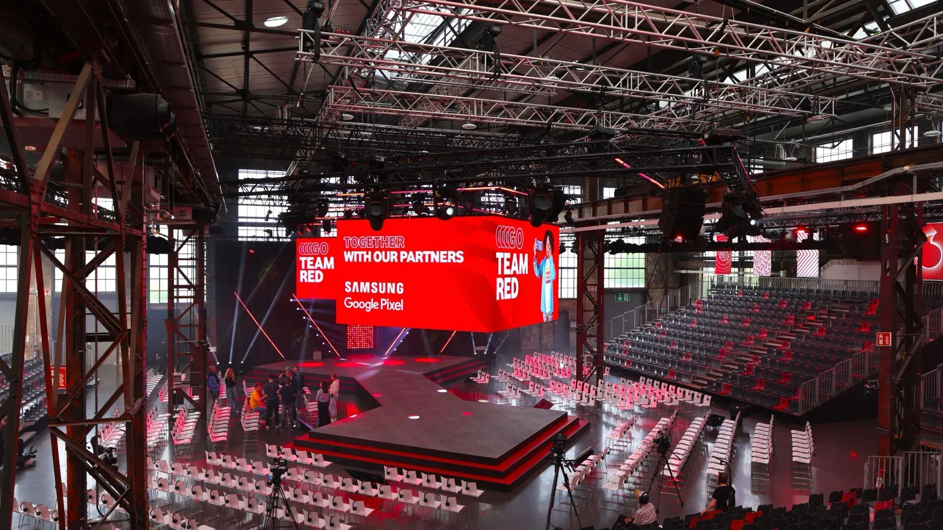 Großes Bühnenbild für eine Veranstaltung mit roter Leinwand, Sitzreihen und Deckenbeleuchtung.