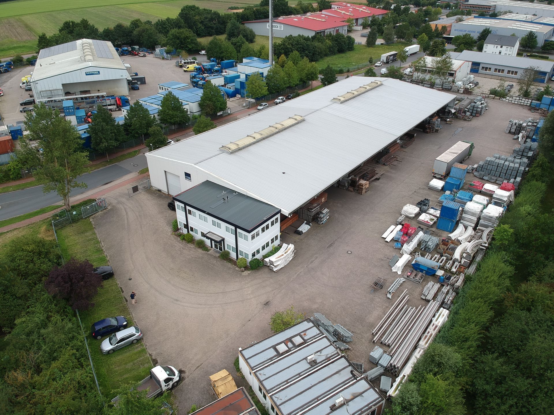 Luftaufnahme eines Industriekomplexes mit Gebäuden, Lagerhallen und geparkten Fahrzeugen in einer grünen Umgebung.