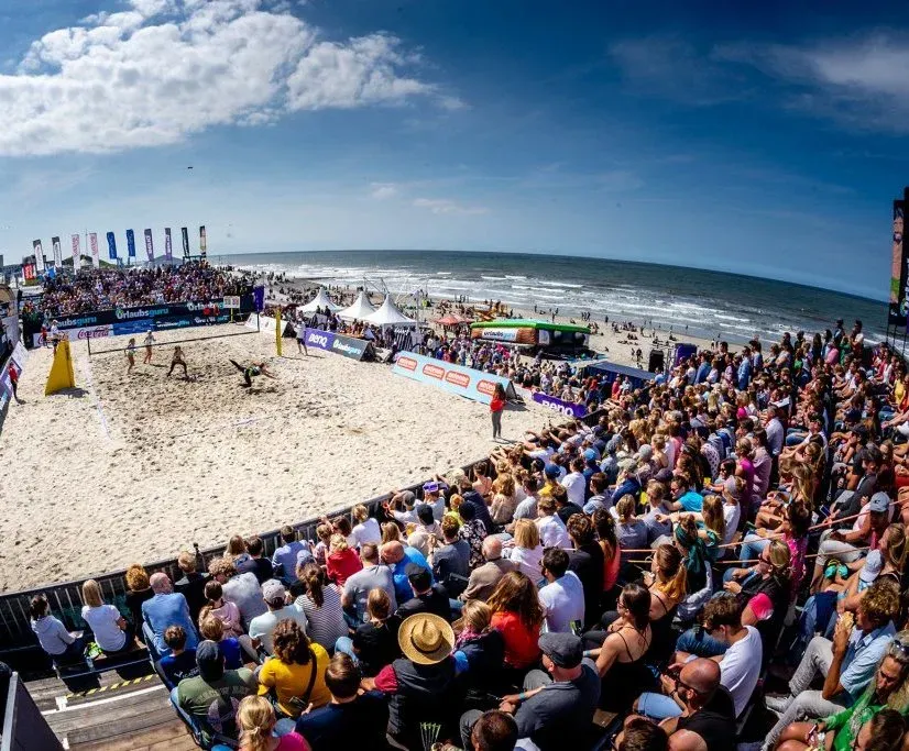 Beachvolleyballspiel mit Zuschauern, sonniger Tag am Meer. Spieler springen, Publikum schaut von der Tribüne aus zu.