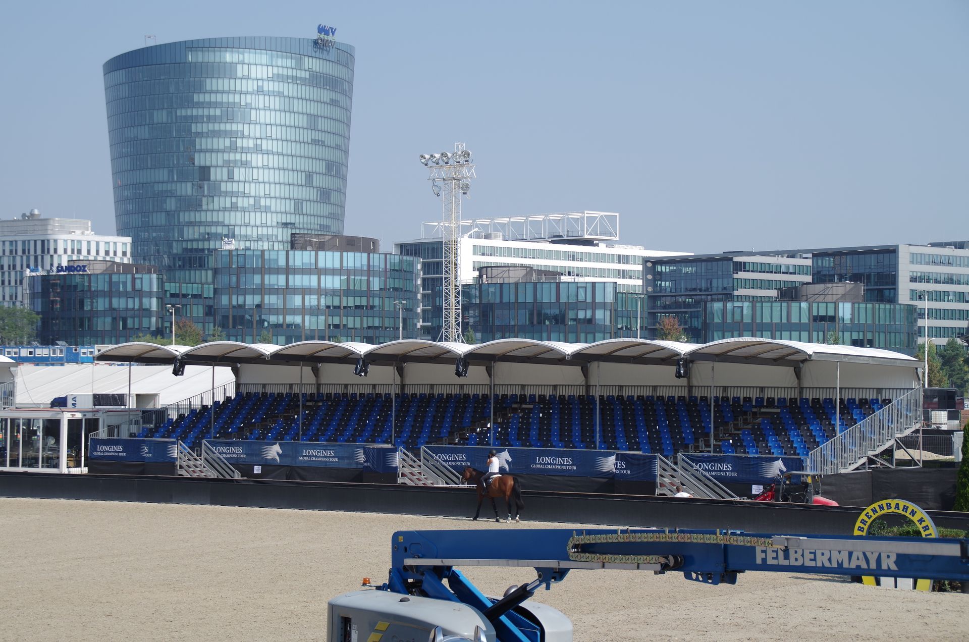 Reitarena mit Stadionbestuhlung, moderne Gebäude im Hintergrund, sonniger Tag.