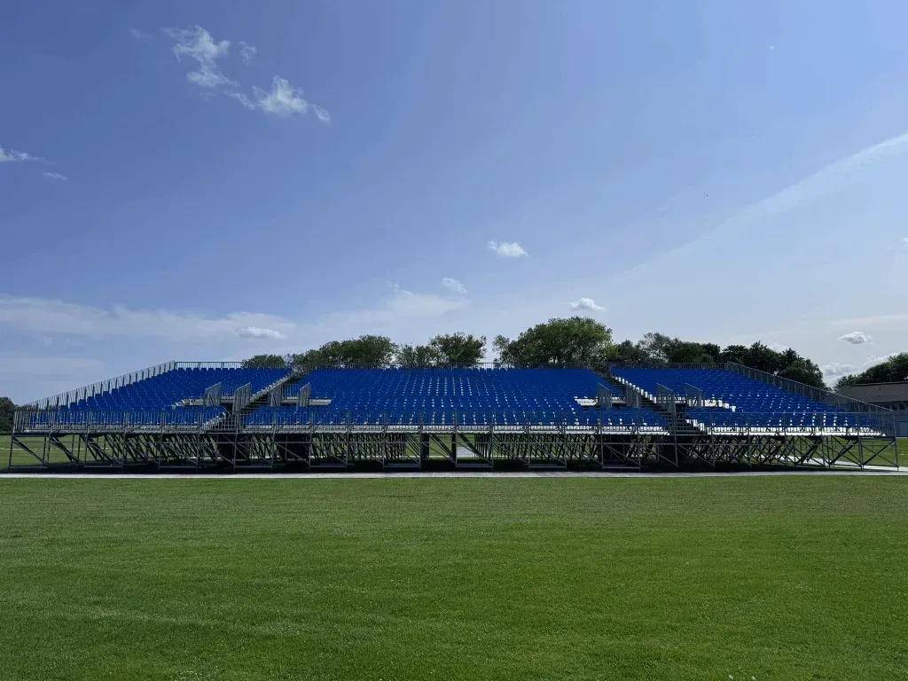 Bleacher seating, blue seats, on a green field under a blue sky.