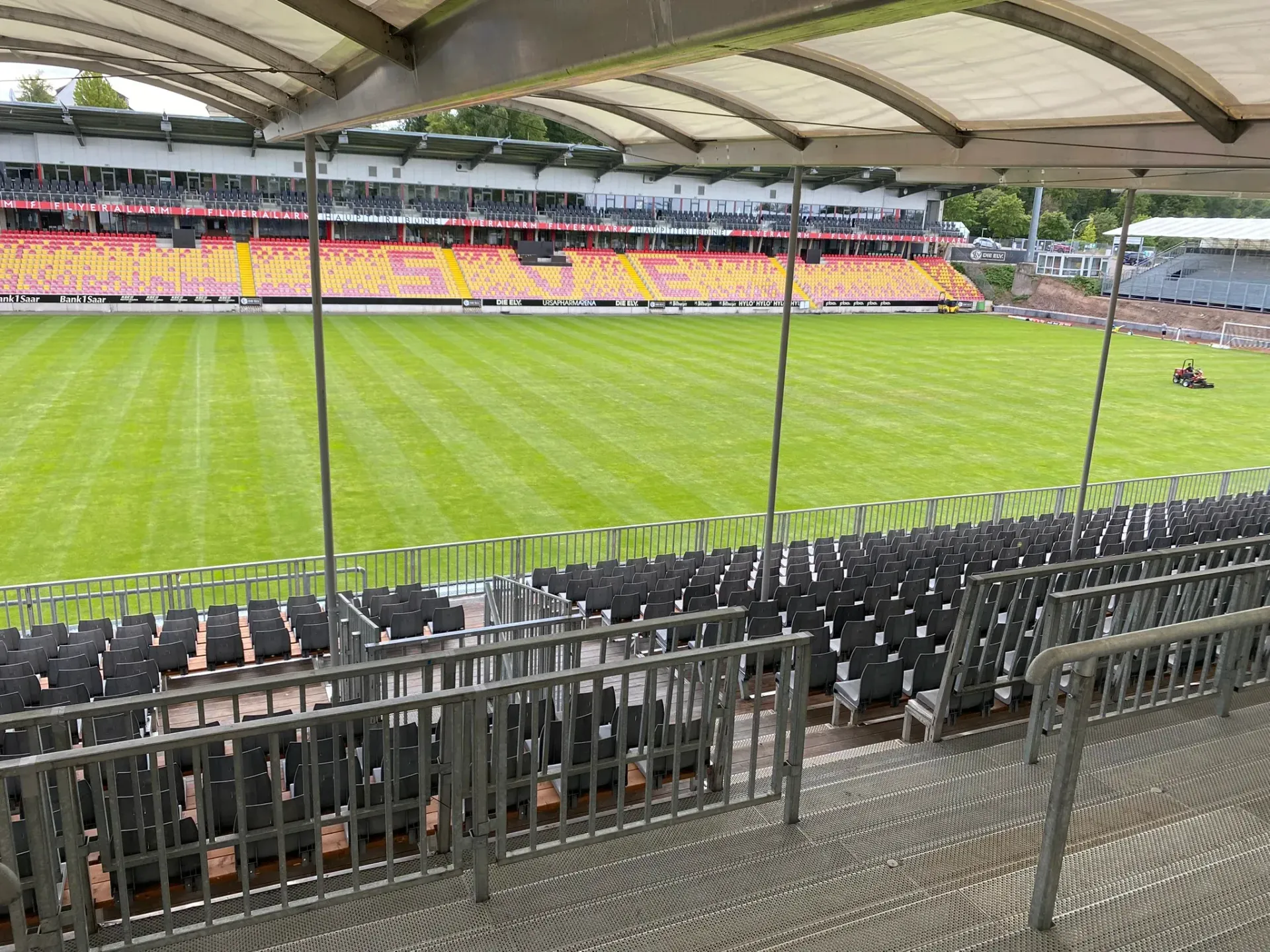 Blick von den Stadionsitzen auf ein grünes Feld. Reihen schwarzer Sitze und gelb/rote Sitze im Hintergrund.