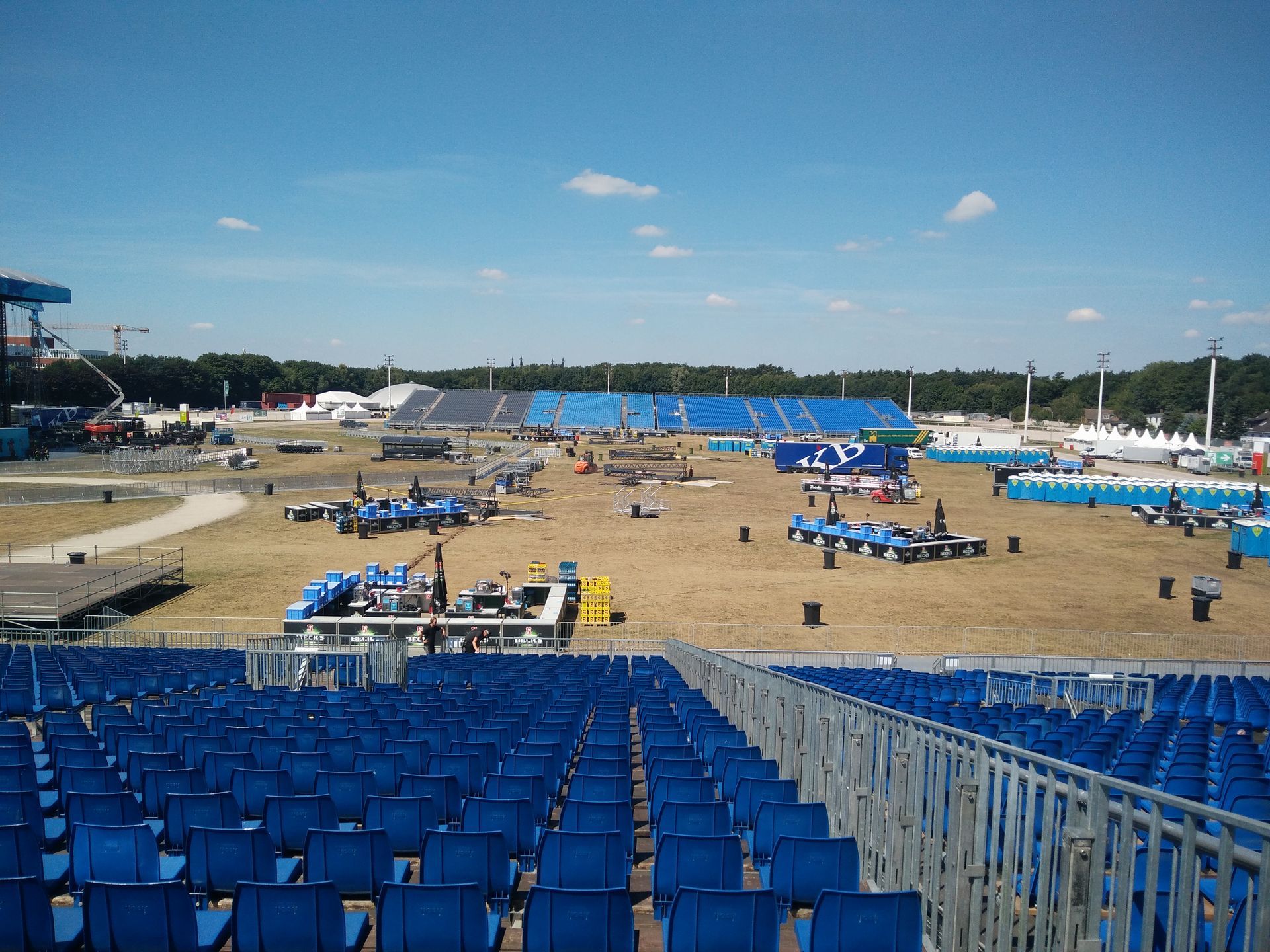 Blue stadium seating overlooking a large outdoor venue with equipment and structures under a blue sky.