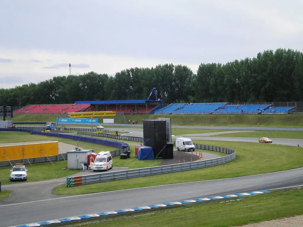Rennwagen auf einer Rennstrecke, im Hintergrund Stadionbestuhlung, Einsatzfahrzeuge.