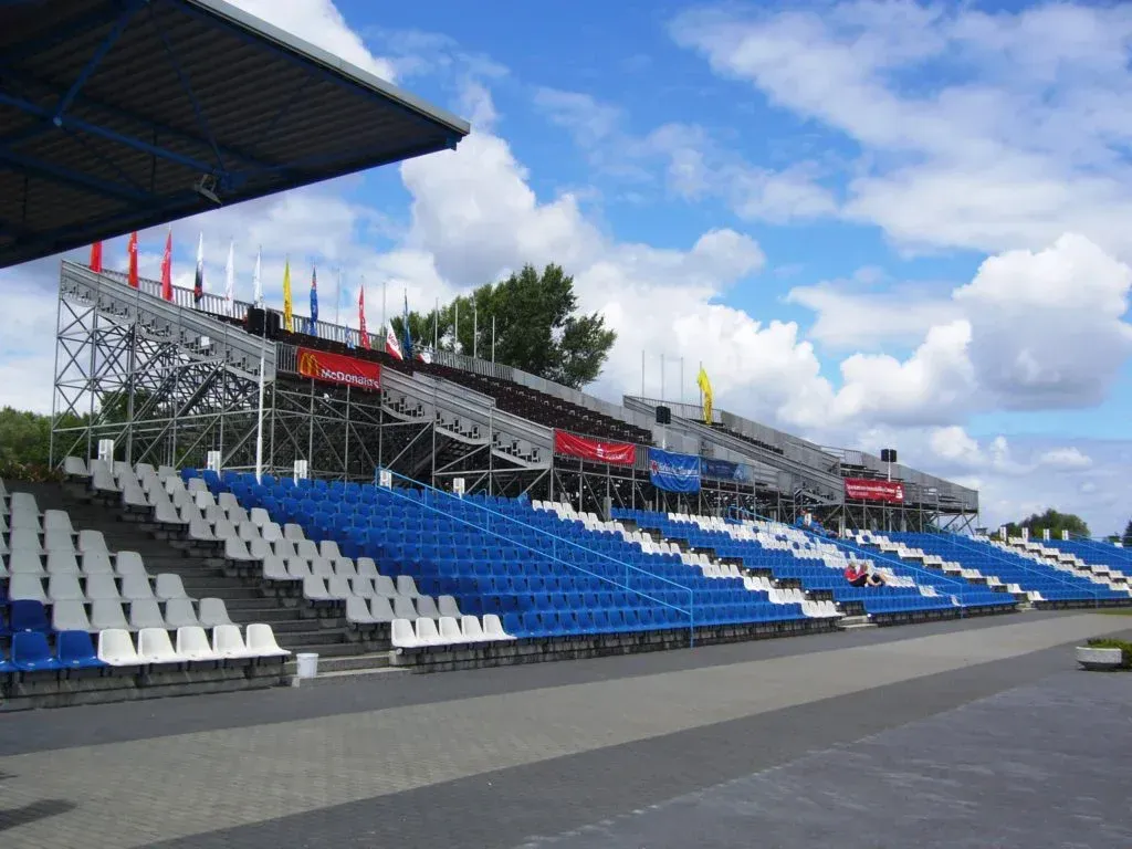 Stadionbestuhlung: blau-weiße Sitze, Metallgerüst, Fahnen vor bewölktem Himmel.