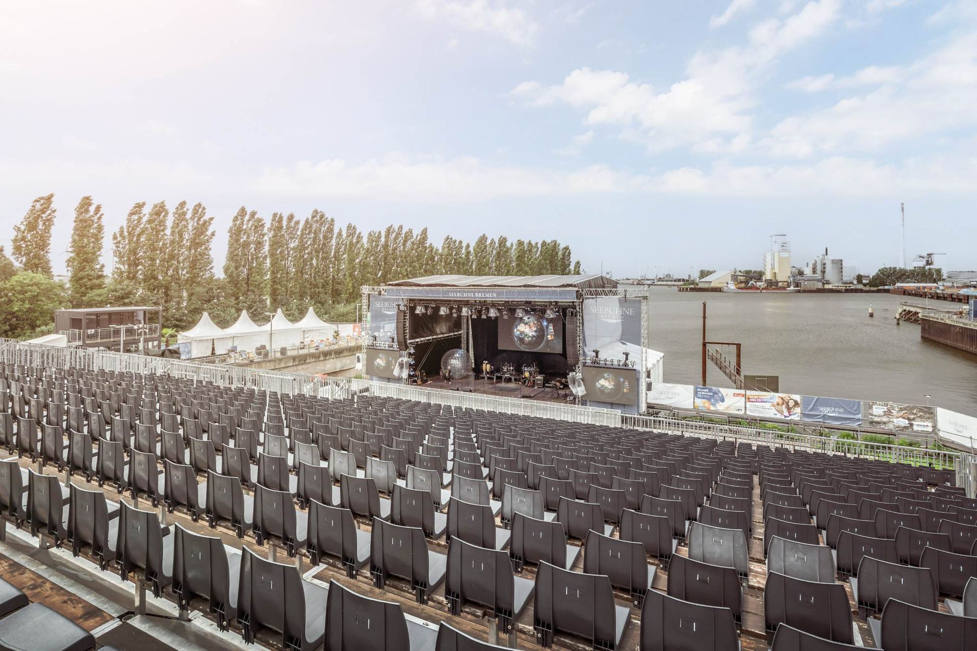 Konzertsaal im Freien mit Bühne, Sitzgelegenheiten und Wasserkulisse unter strahlendem Himmel.