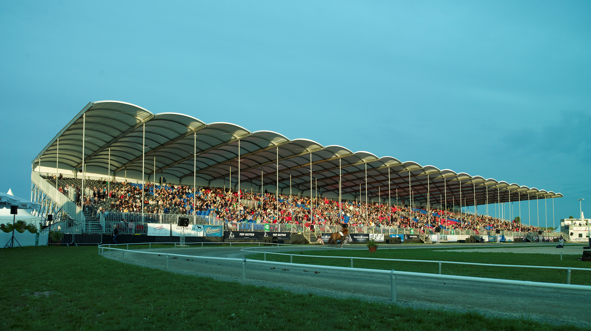 Unter einem weißen Dach sind die Zuschauerränge mit Menschen gefüllt. Grünes Gras und blauer Himmel sind zu sehen.