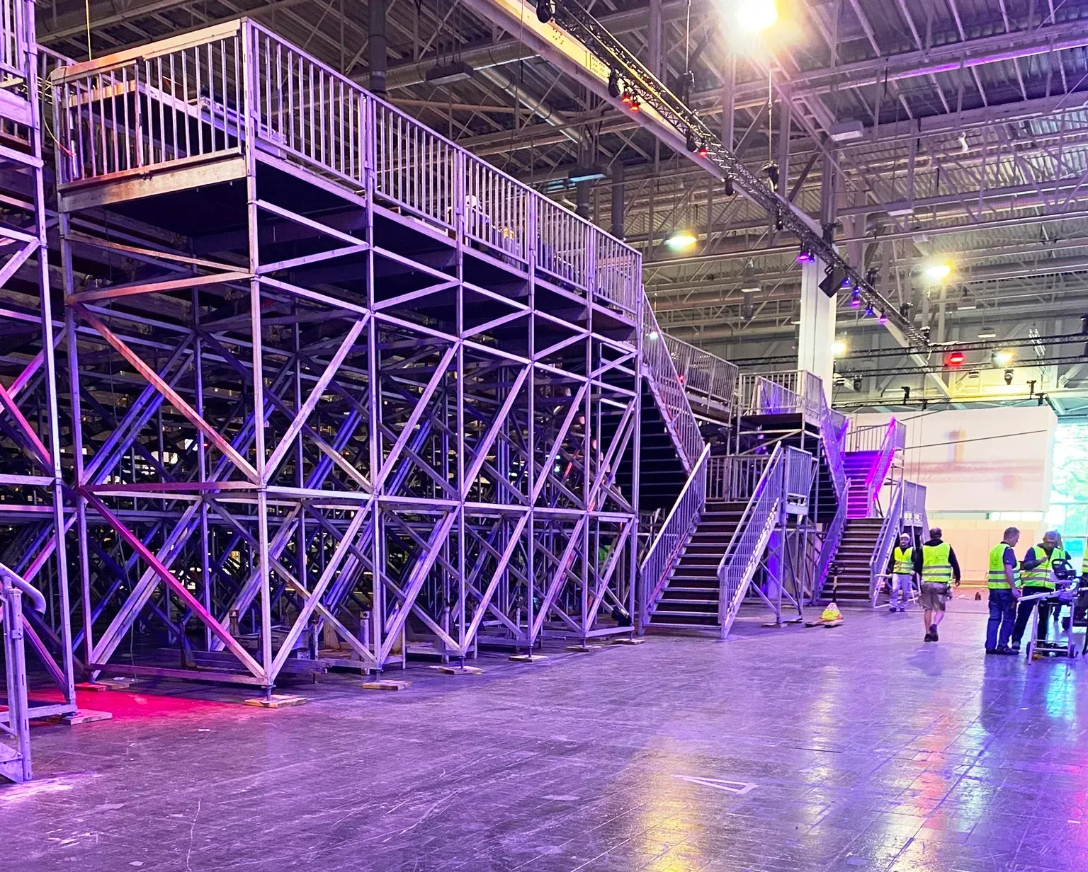 Metallgerüstkonstruktion in einer großen Halle mit Treppen und einer erhöhten Plattform. Arbeiter in Westen sind zu sehen.
