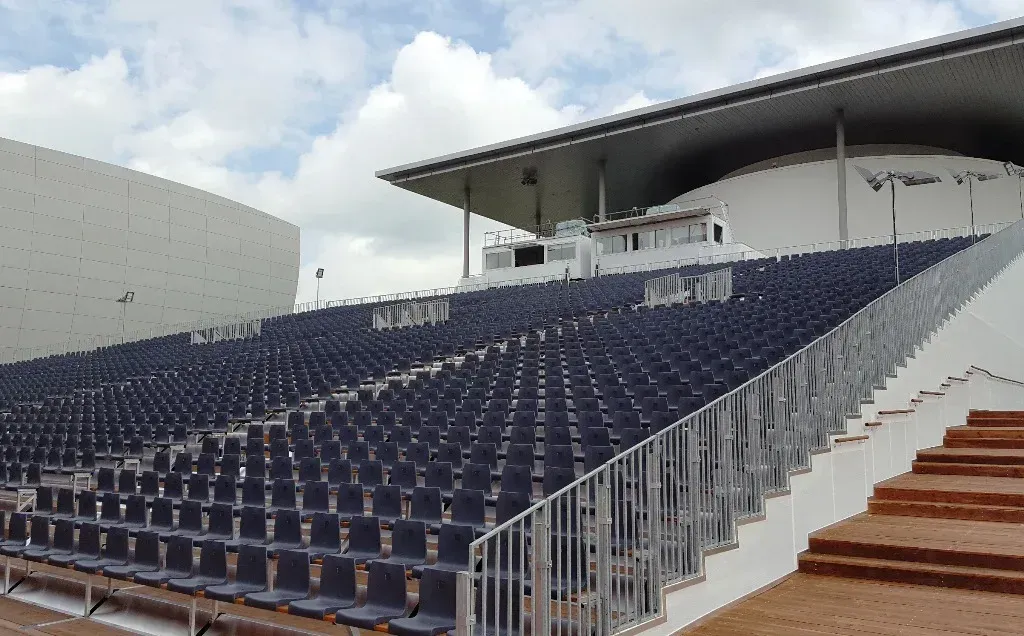 Reihen blauer Stadionsitze unter einem weißen Baldachin; im Hintergrund bewölkter Himmel.