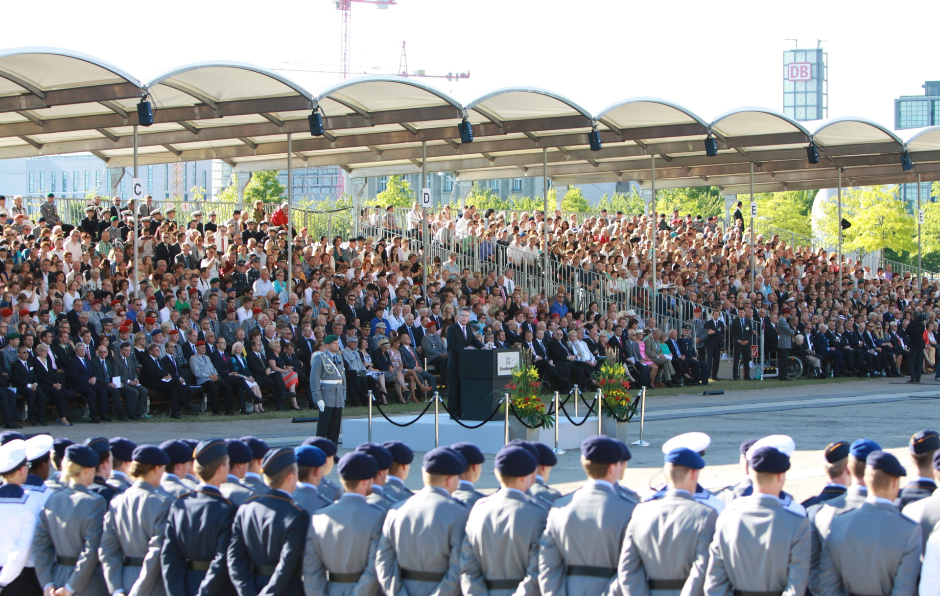 Zeremonie mit einem Redner auf einem Podium, Militärpersonal in Formation und einer großen Menschenmenge auf den Tribünen im Freien.
