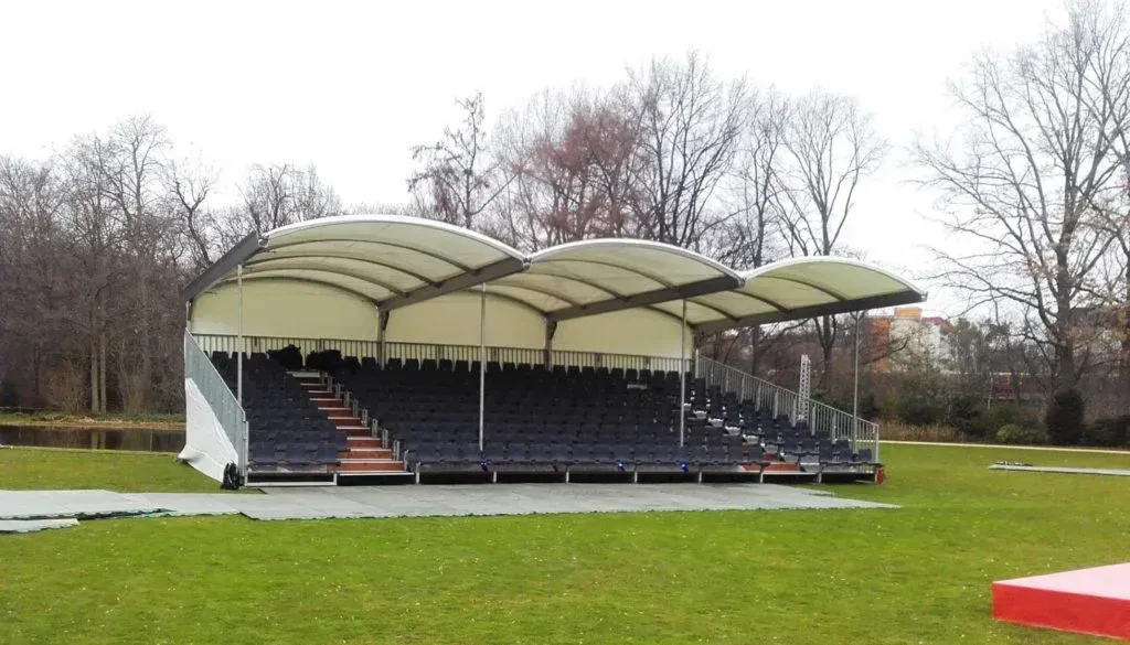 Freiluftstadion mit Reihen blauer Sitze unter einem weißen Baldachin, in einem Park mit Bäumen gelegen.