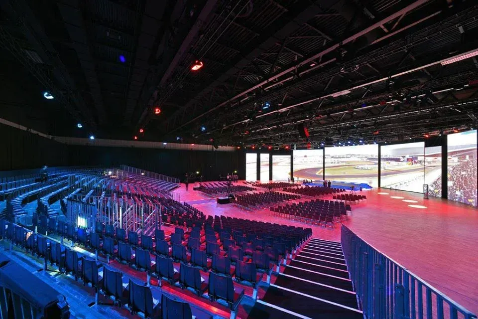 Großes Theater mit Sitzreihen, Bühne und großen Bildschirmen; blaue und rote Beleuchtung.