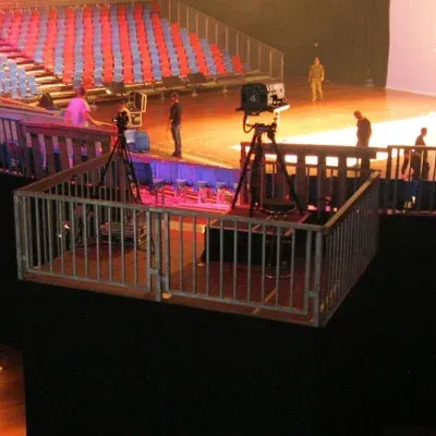 Kameras auf einer umzäunten Plattform bei einer Veranstaltung, mit Menschen und Bühnensitzen im Hintergrund.