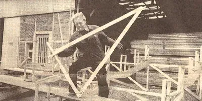 Mann baut in einer Werkstatt einen Holzrahmen, im Hintergrund ist gestapeltes Bauholz zu sehen.