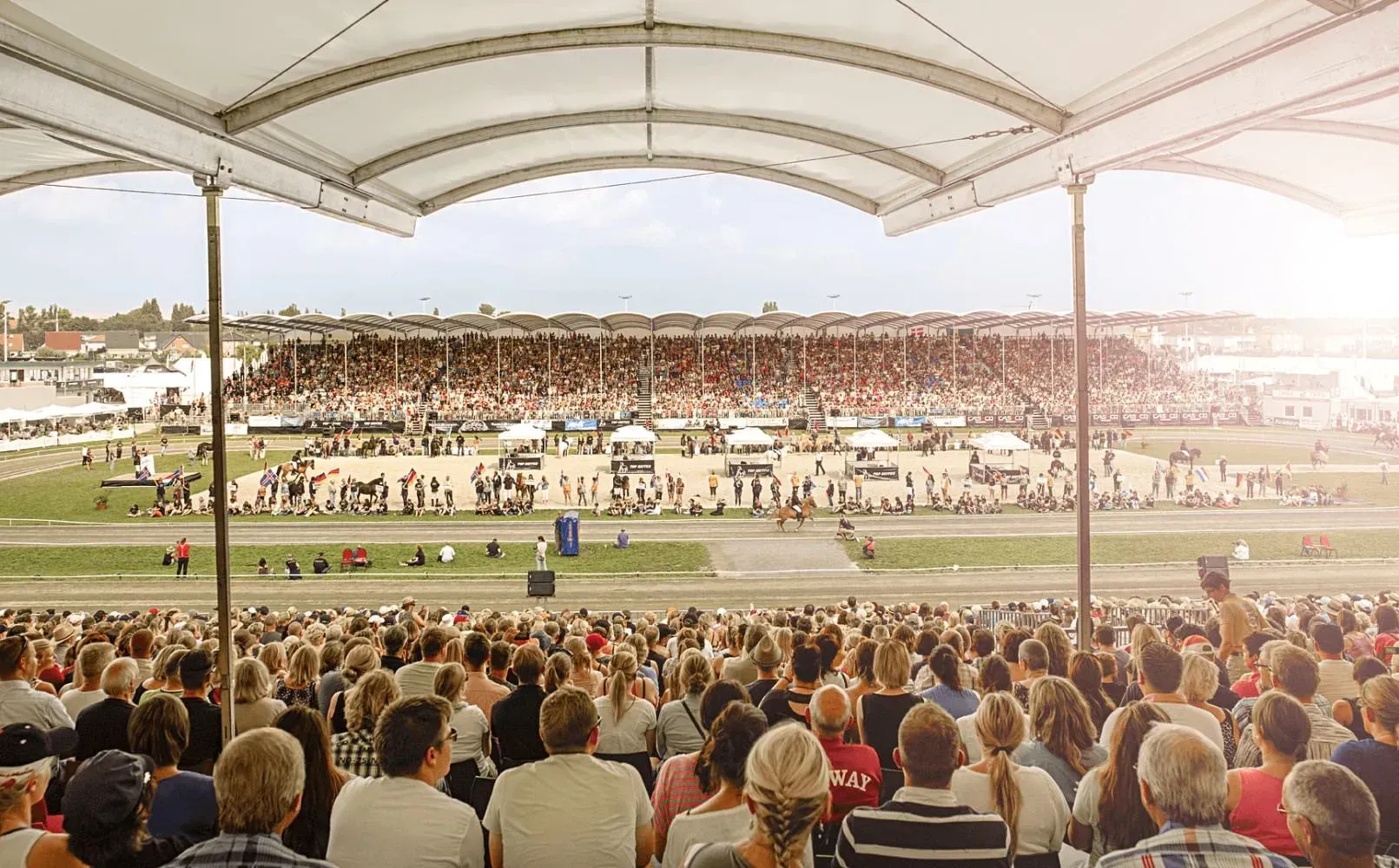 Zuschauer verfolgen ein Reitturnier im Freien unter einer überdachten Tribüne. Ein sonniger Tag mit vielen Menschen.