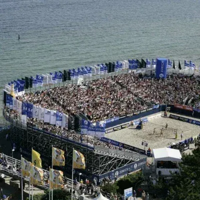 Beachvolleyballstadion mit vollen Tribünen am Meer. Spieler auf dem Platz. Blau-weißes Branding.