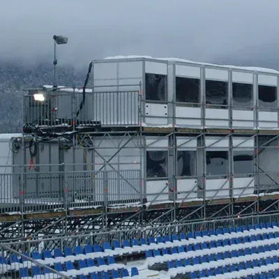 Zweistöckiges, schneebedecktes Gebäude auf den Sitzplätzen des Stadions; im Hintergrund schneebedeckte Berge.