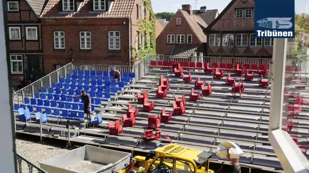 Blaue und rote Sitze in einem gestuften Freilufttheater, Baumaschinen, historische Gebäude im Hintergrund.