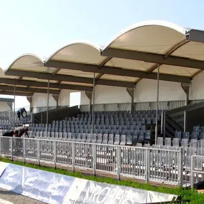 Sitzplätze im Stadion im Freien unter einem geschwungenen weißen Baldachin; graue Stühle, Metallgeländer.