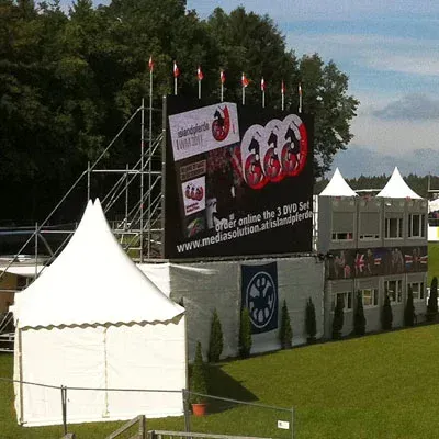 Weiße Zelte und ein modulares Gebäude mit einer großen Leinwand und Flaggen auf einer Wiese.