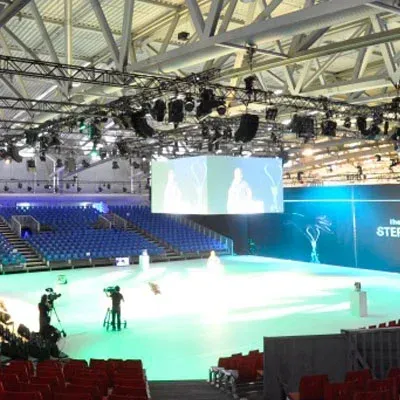 Eine leere Arena mit Bühne, großer Leinwand und blauen Sitzen. Das Bühnenteam filmt eine Präsentation vor hellgrünem Boden.