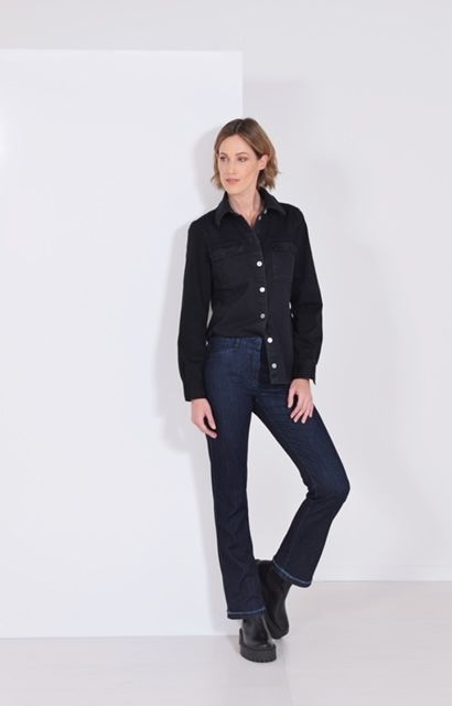 Eine Frau in einem schwarzen Hemd und blauen Jeans steht vor einer weissen Wand.