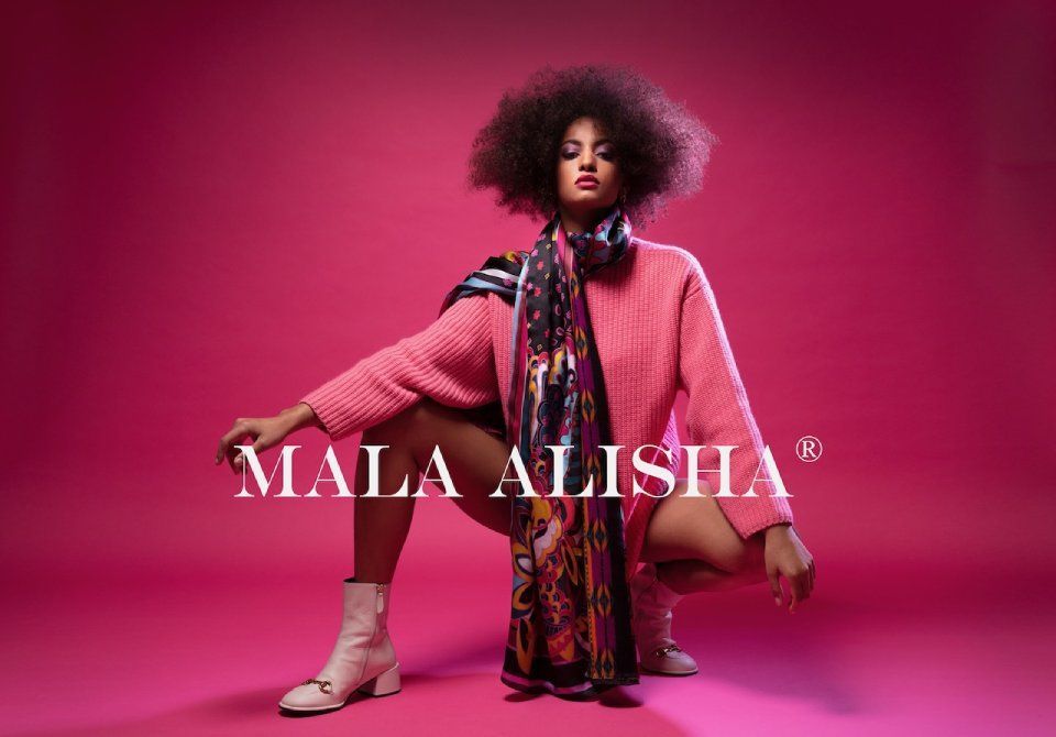 Eine Frau in einem rosa Pullover und Schal kniet vor einem rosa Hintergrund. Foto von Mala Alisha