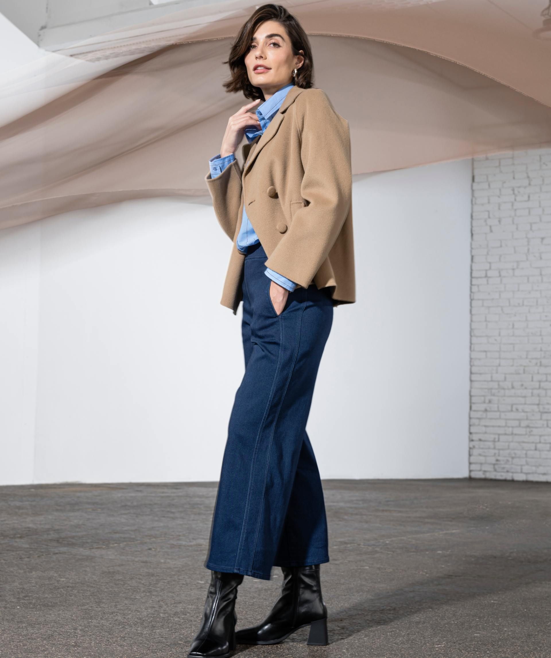 Eine Frau in einer hellbraunen Jacke und blauen Hosen steht vor einer weissen Wand.