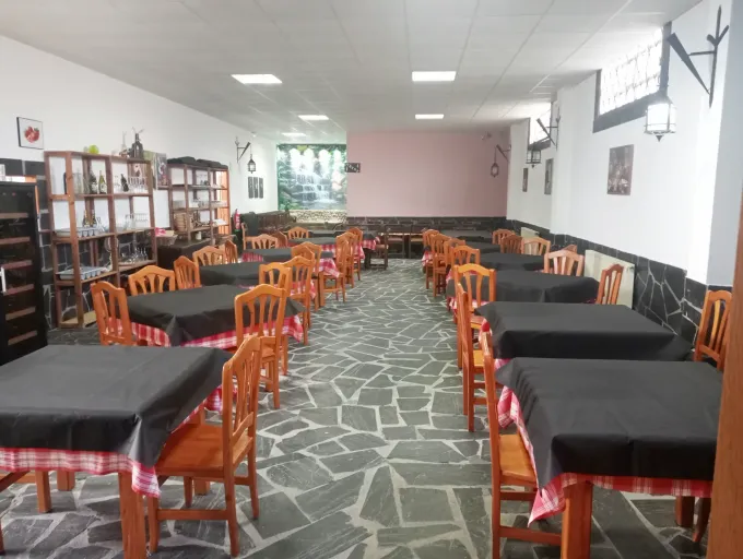 Interior de restaurante con mesas, sillas y suelo de piedra. Paredes blancas con molduras oscuras y ventanas arqueadas.