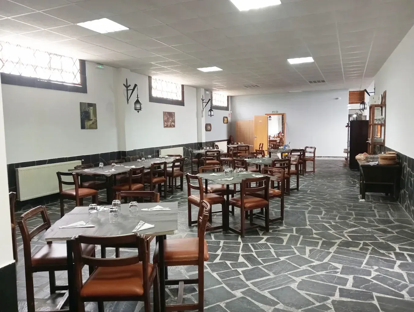 Interior de restaurante con mesas, sillas y suelo de piedra. Paredes blancas con molduras oscuras y ventanas arqueadas.