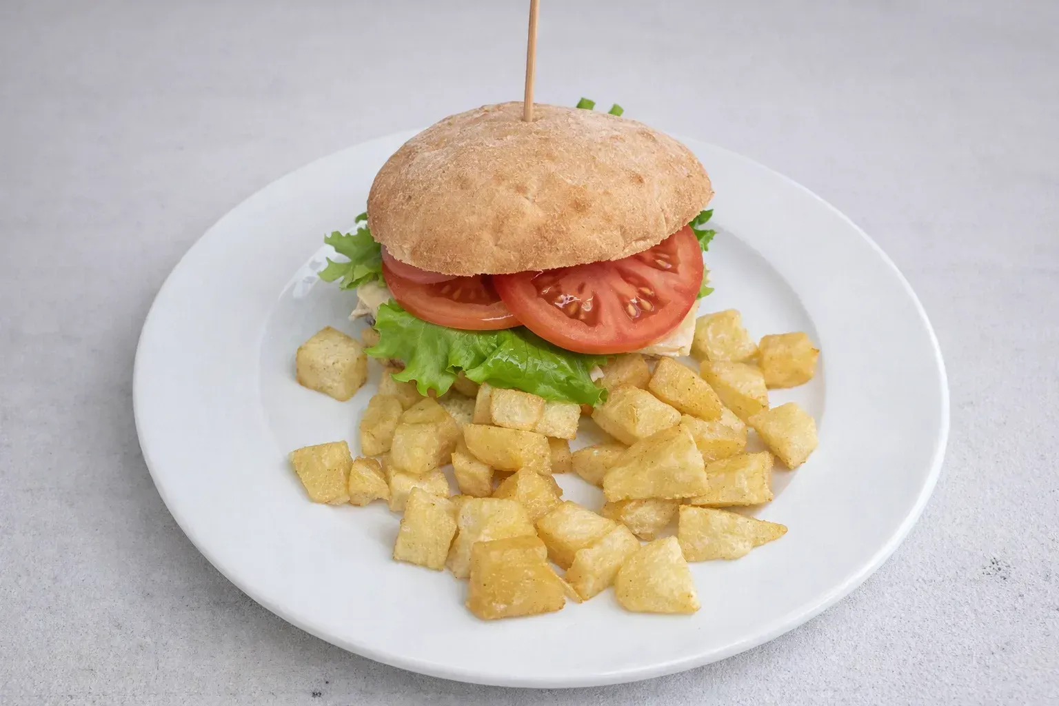 Sándwich en pan con lechuga y tomate, ensartado con un palillo, servido con patatas fritas en un plato blanco.