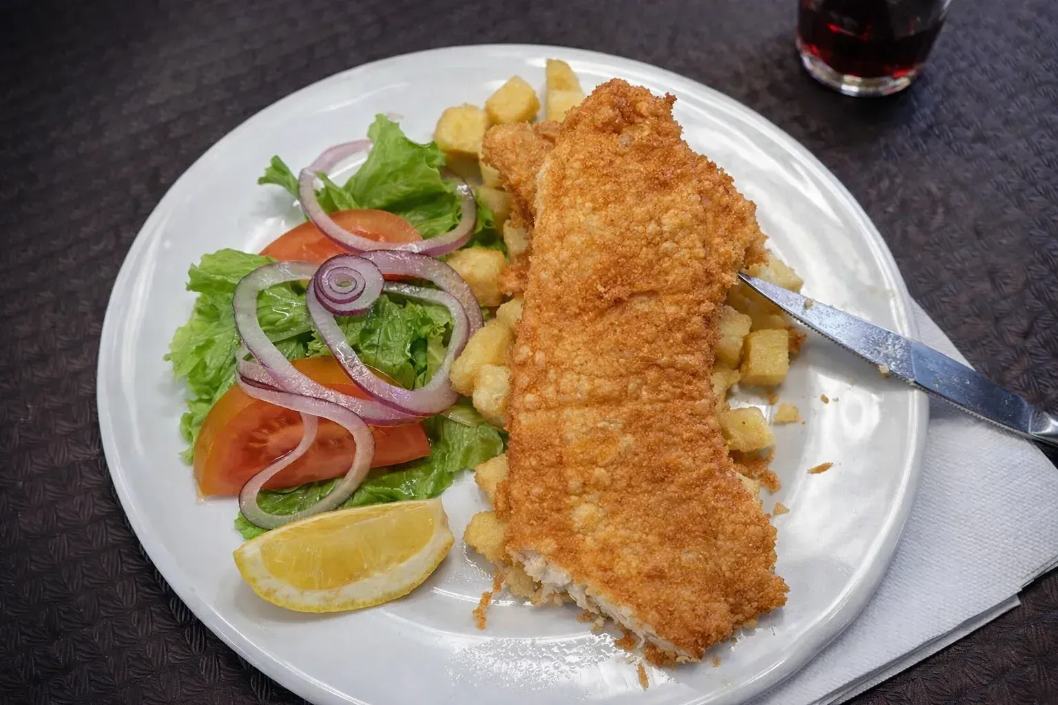 Pescado empanizado con patatas fritas, ensalada y limón en un plato blanco.