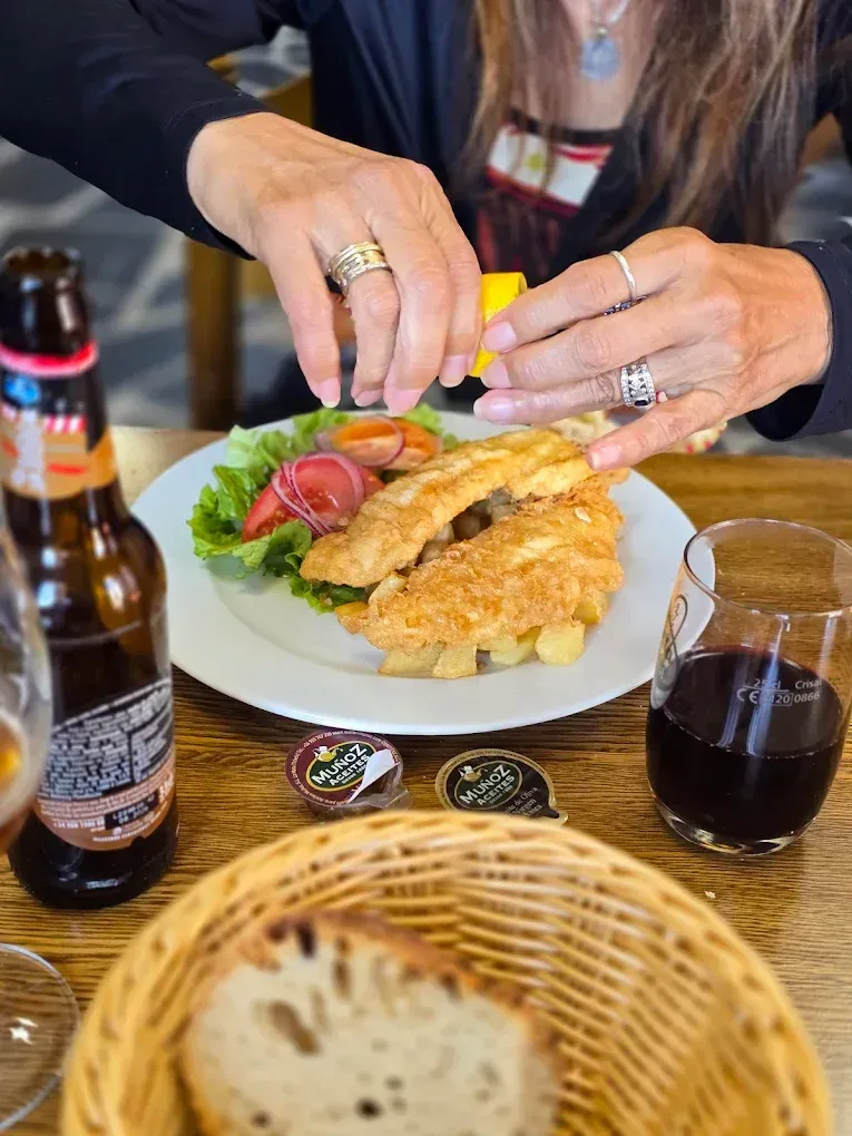 Persona exprimiendo limón sobre pescado frito y patatas fritas en un plato. Cerveza, vino y cesta de pan a la vista.