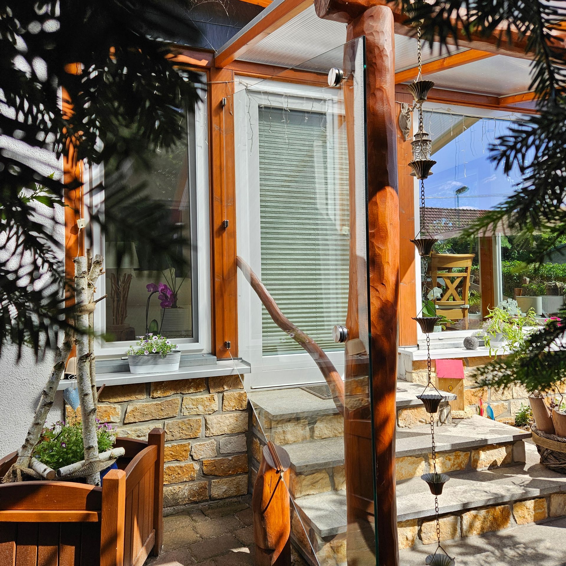Veranda mit Steinstufen, orangefarbenen Holzpfosten, Glastür und Topfpflanzen im hellen Sonnenlicht
