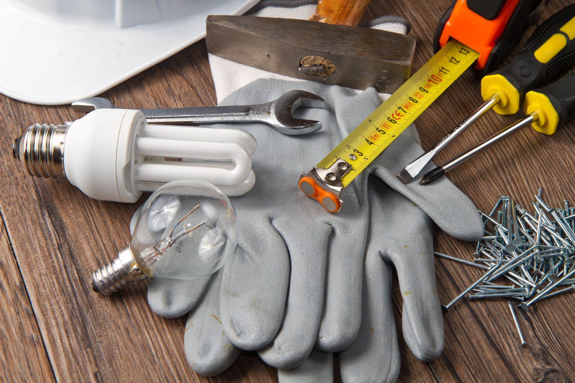 Outils et fournitures sur une surface en bois : gants, ampoule, clé, marteau, mètre ruban, clous et tournevis.