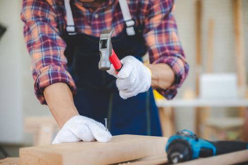 Une personne enfonce un clou dans une planche de bois dans un atelier, portant des gants et une chemise à carreaux.