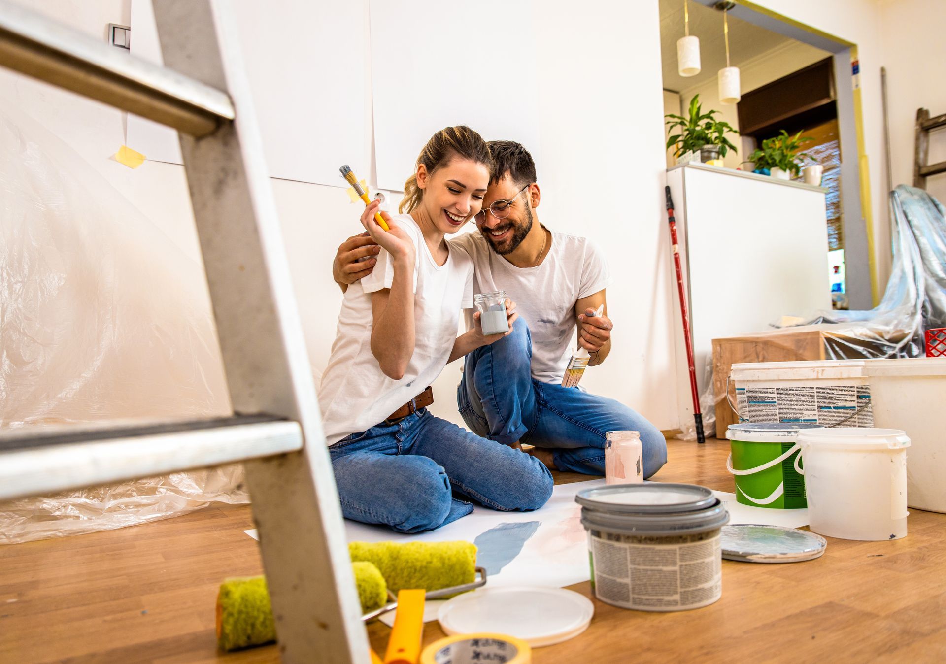 Un couple peint un mur à l'intérieur, souriant assis par terre avec des pinceaux en main.