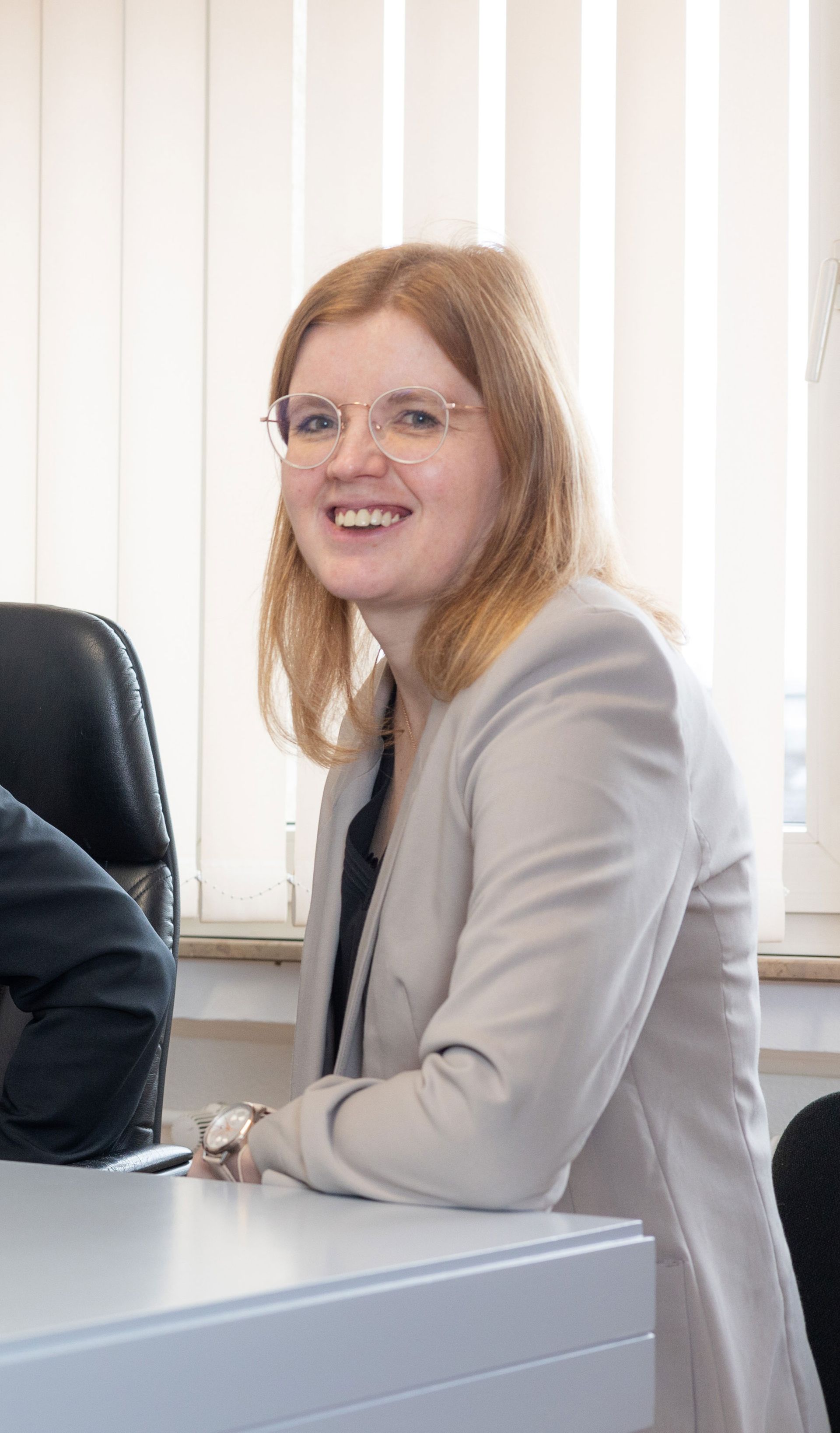 Eine Frau in Anzug und Brille lehnt an einem Schreibtisch und lächelt.