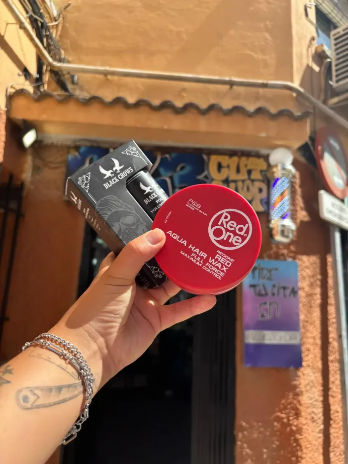 Persona sosteniendo un producto para peinar el cabello Red One y un producto Black Dragon afuera de una peluquería.