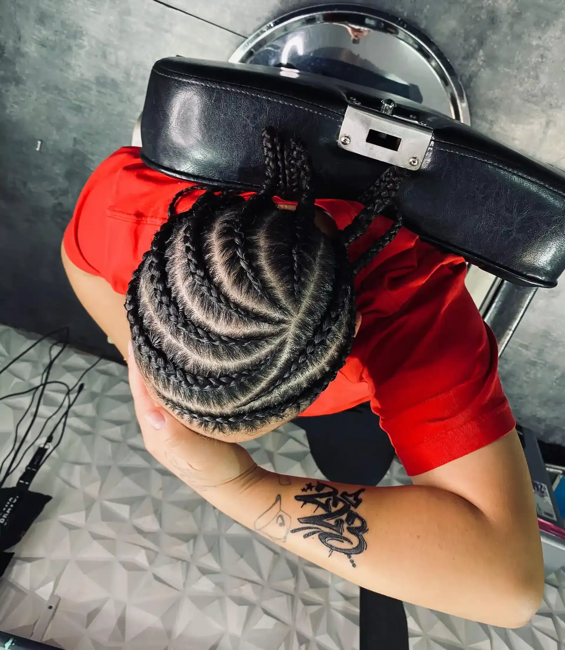 Persona con cabello trenzado en espiral, con camisa roja, sentada en una silla de salón. Brazo con tatuaje visible.