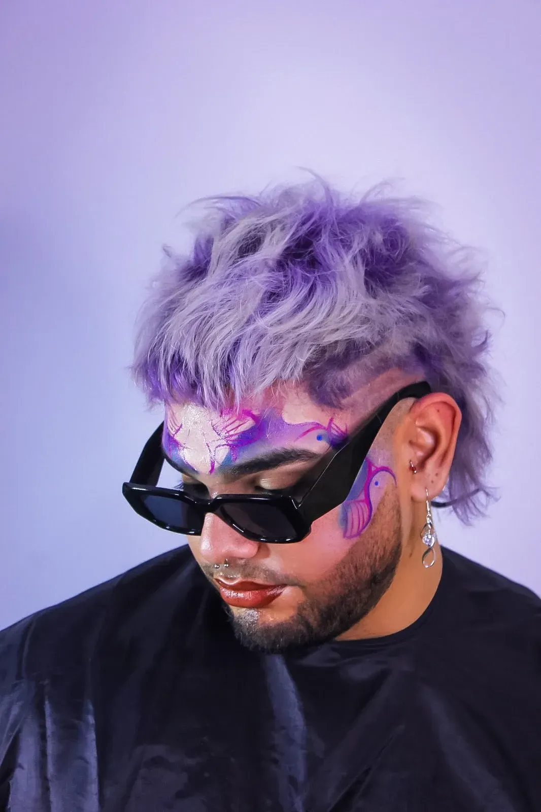 Persona con cabello morado, gafas de sol y pintura facial. Vestida de negro, mirando hacia abajo.