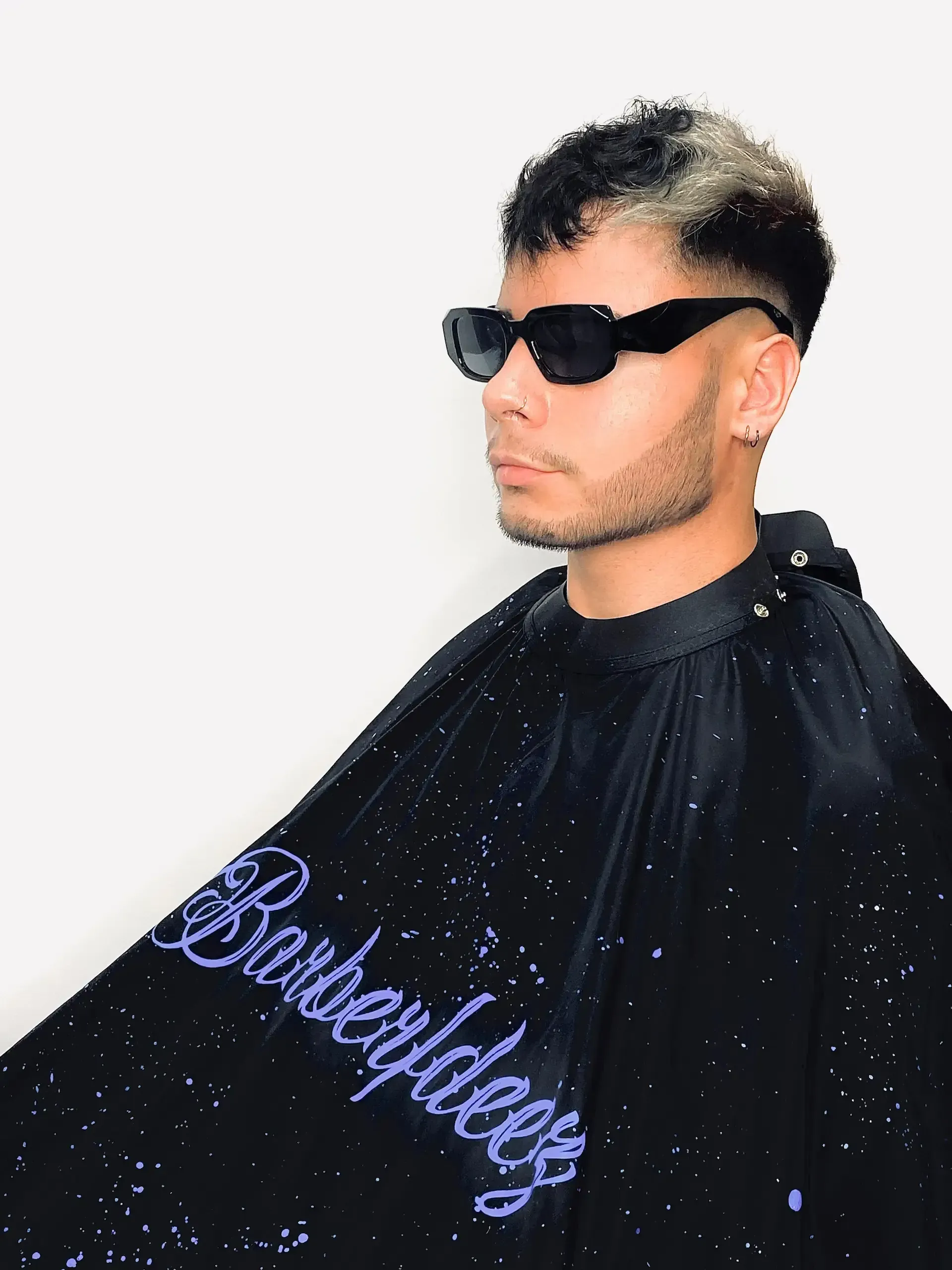 Hombre con gafas de sol y capa de barbero, con cabello claro y un lado afeitado.