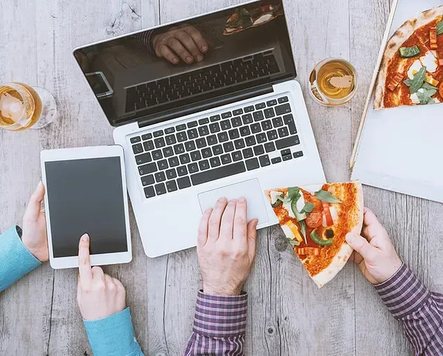 Un hombre está comiendo una porción de pizza mientras usa una computadora portátil y una tableta.