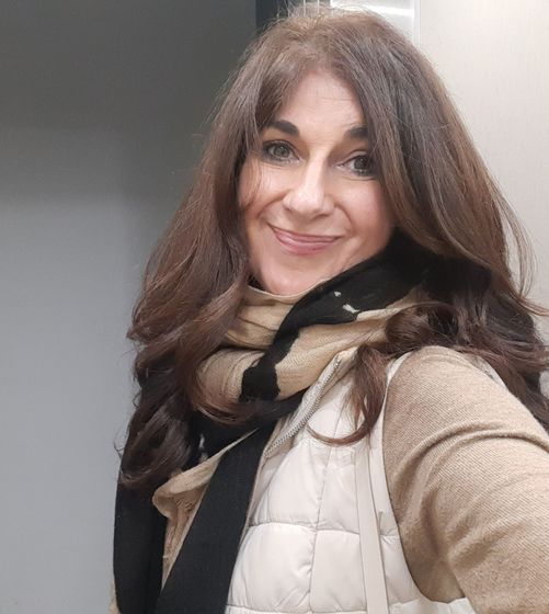 Una persona de cabello castaño sonríe, vistiendo un suéter color canela, una bufanda beige y negra