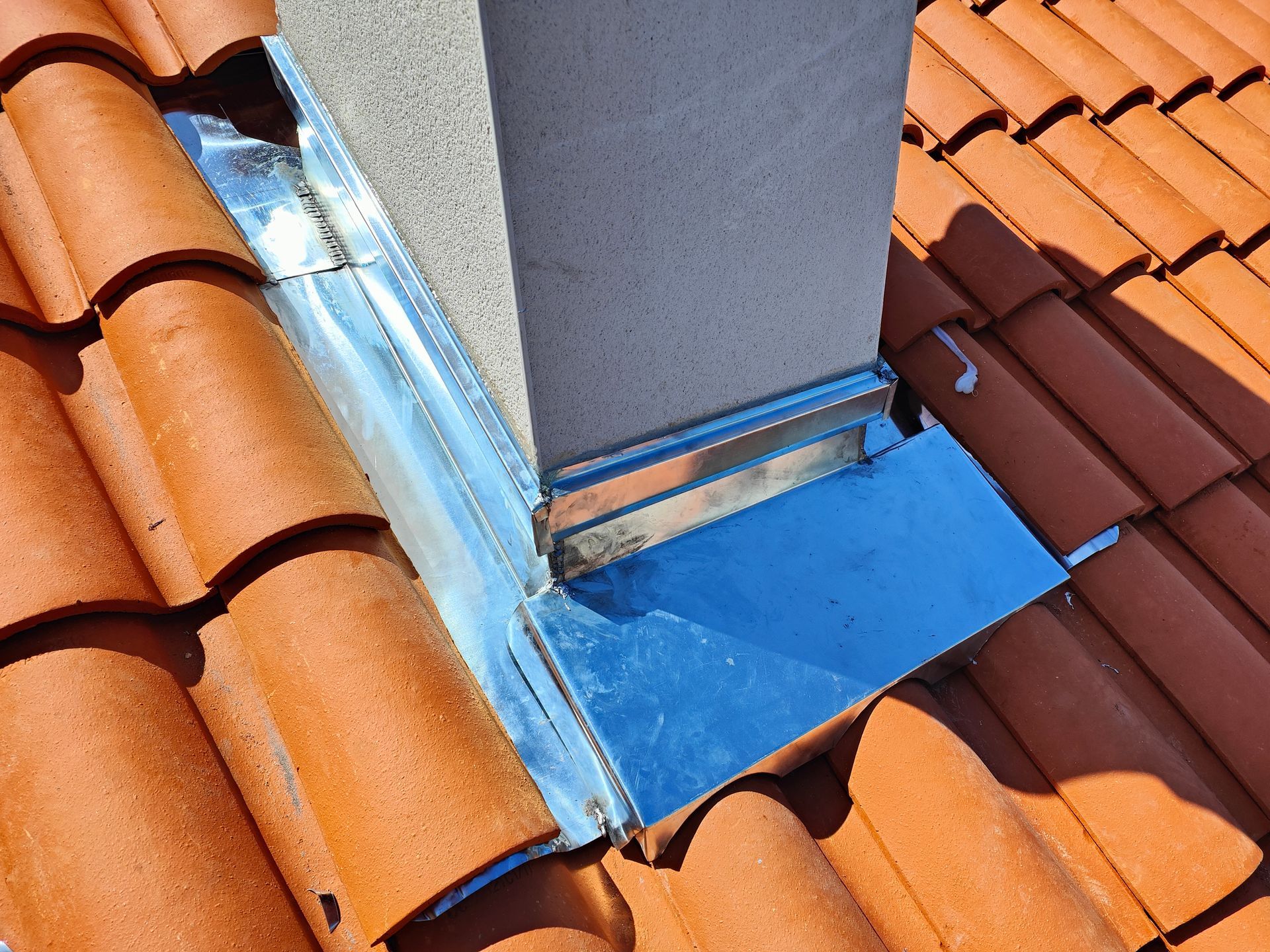 Noue de toit avec liseré bleu entre les tuiles orange et rouges autour d'une cheminée grise.