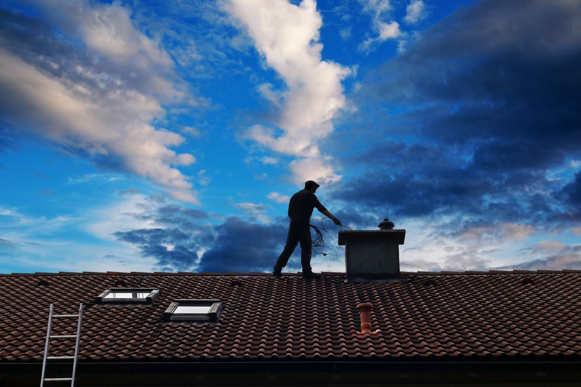 Un ramoneur sur un toit de tuiles, se détachant sur un ciel d'un bleu spectaculaire, utilise un outil près d'une cheminée.