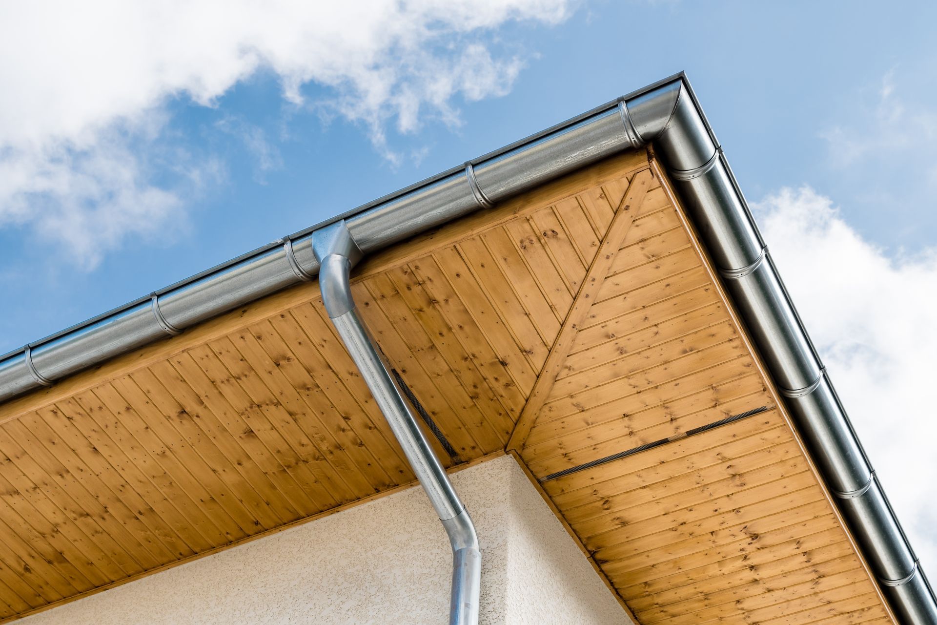 Gros plan sur un avant-toit de maison avec des gouttières grises se détachant sur un ciel bleu et des nuages.