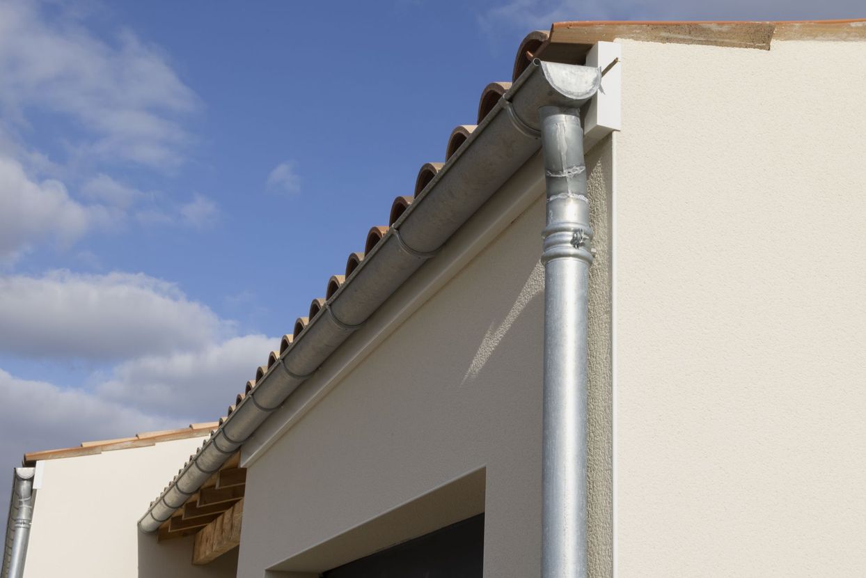 Gros plan sur le bord d'un toit de maison avec une gouttière et une descente pluviale grises sur fond de ciel bleu.