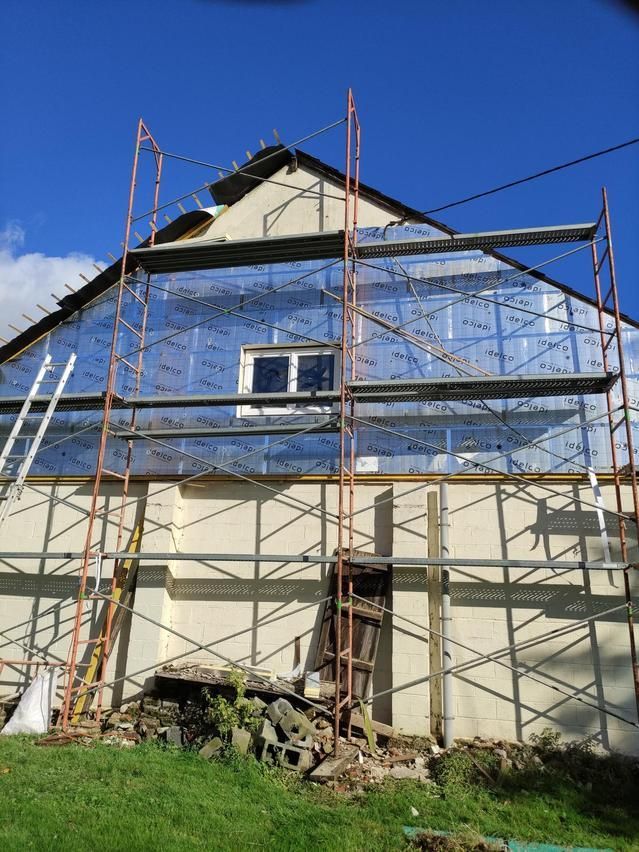 Façade de maison en rénovation, avec échafaudages et pare-vapeur bleu sur fond de ciel dégagé.